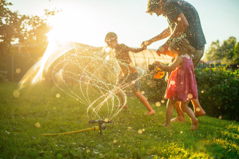 A photo with a sprinkler in the foreground and a parent holding hands with 2 children dancing through the sprinkler. The sun is setting and shining brightly.